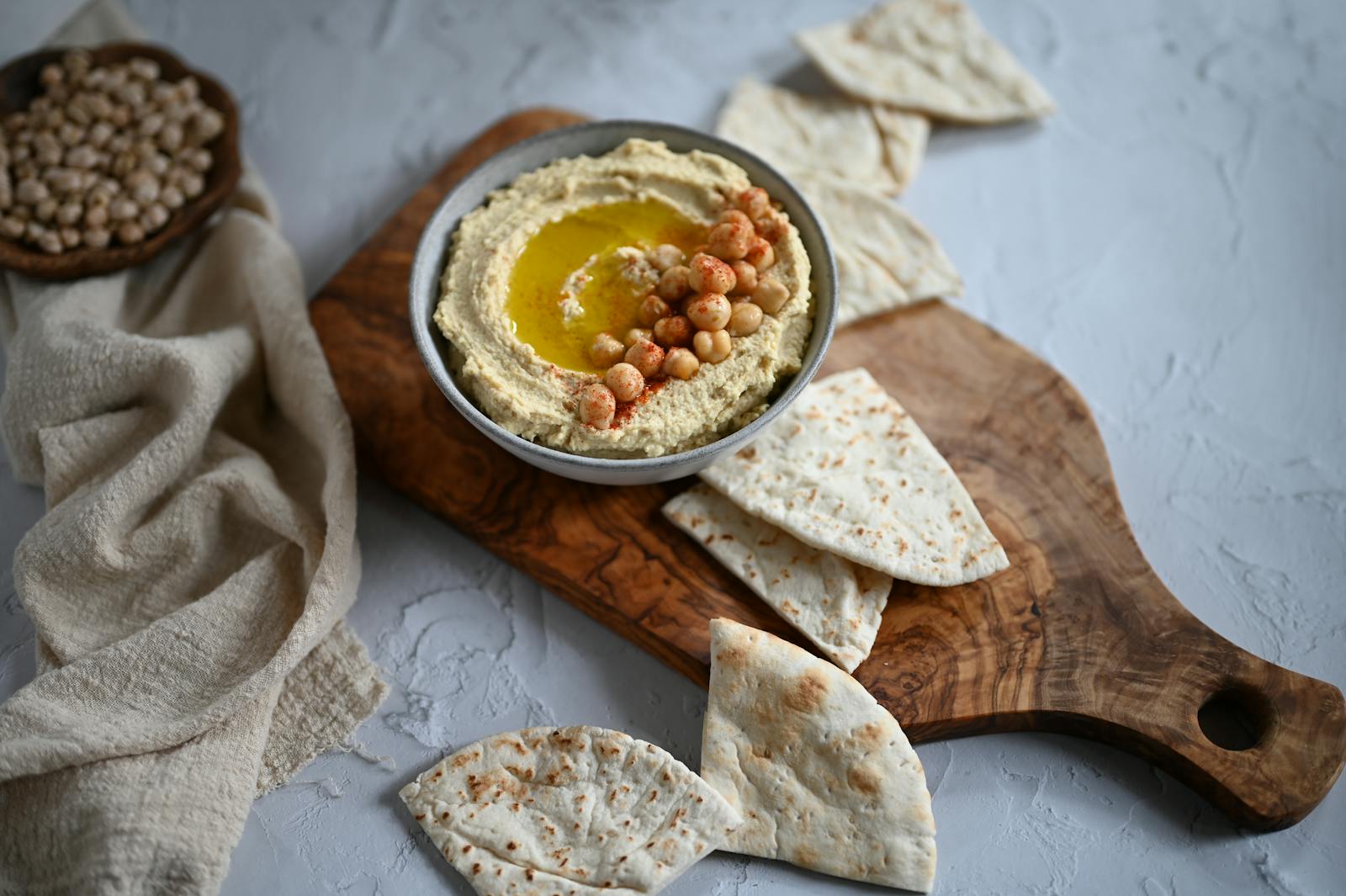 A bowl of creamy hummus garnished with chickpeas and olive oil, served with pita bread on a wooden cutting board.